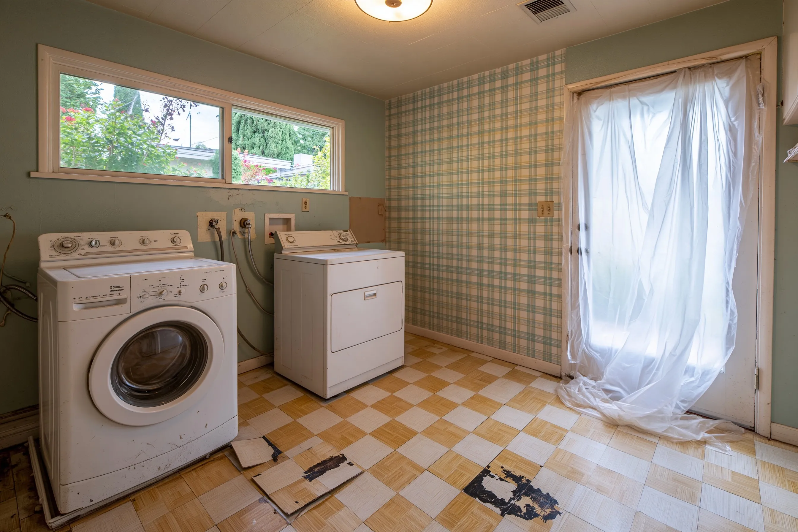 Laundry room with a washer and dryer pulled from the wall, checkered floor tiles lifted and broken over black adhesive, and plastic over the back door